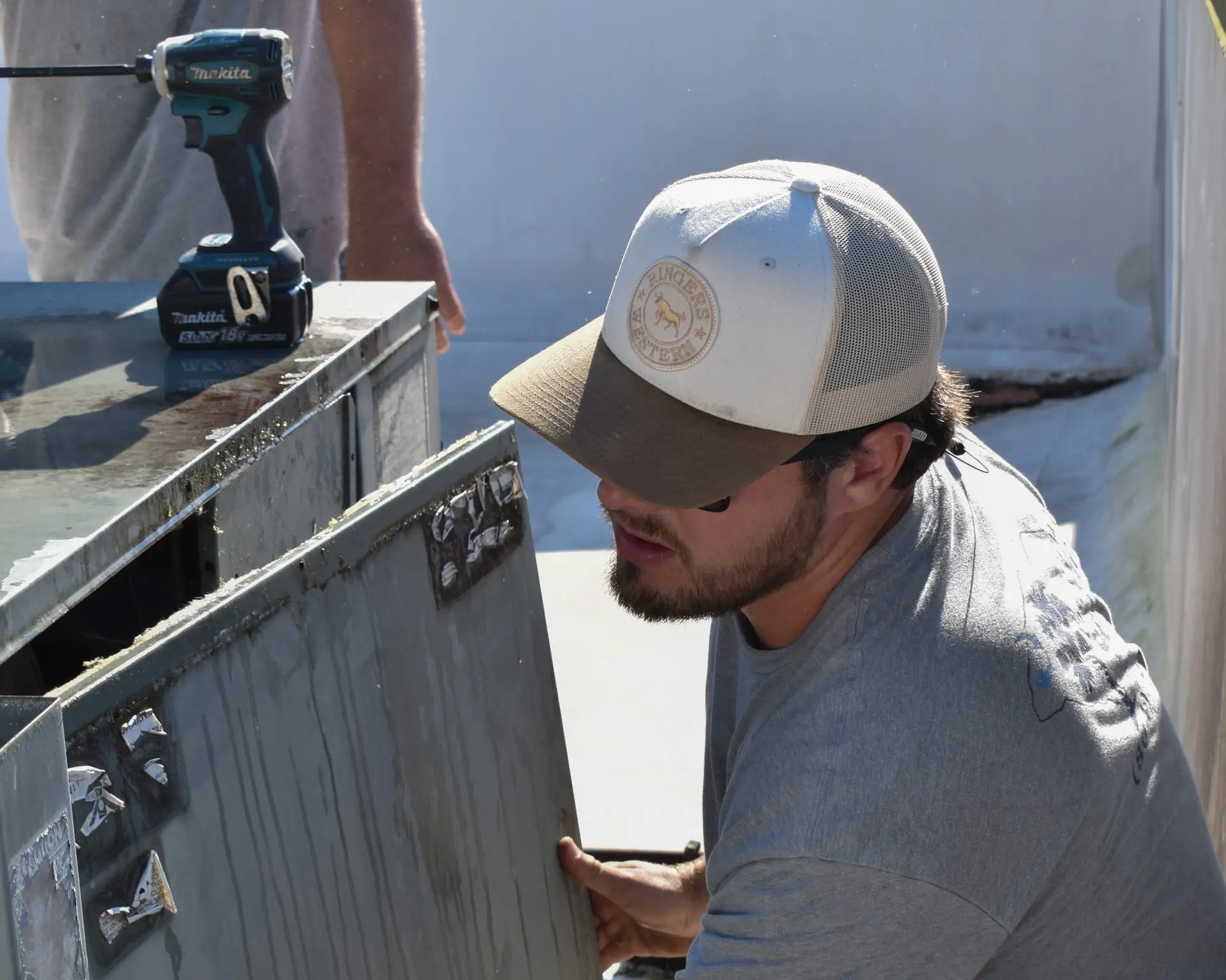 HVAC employee working on HVAC system with hat on and drill on top of system - person in background