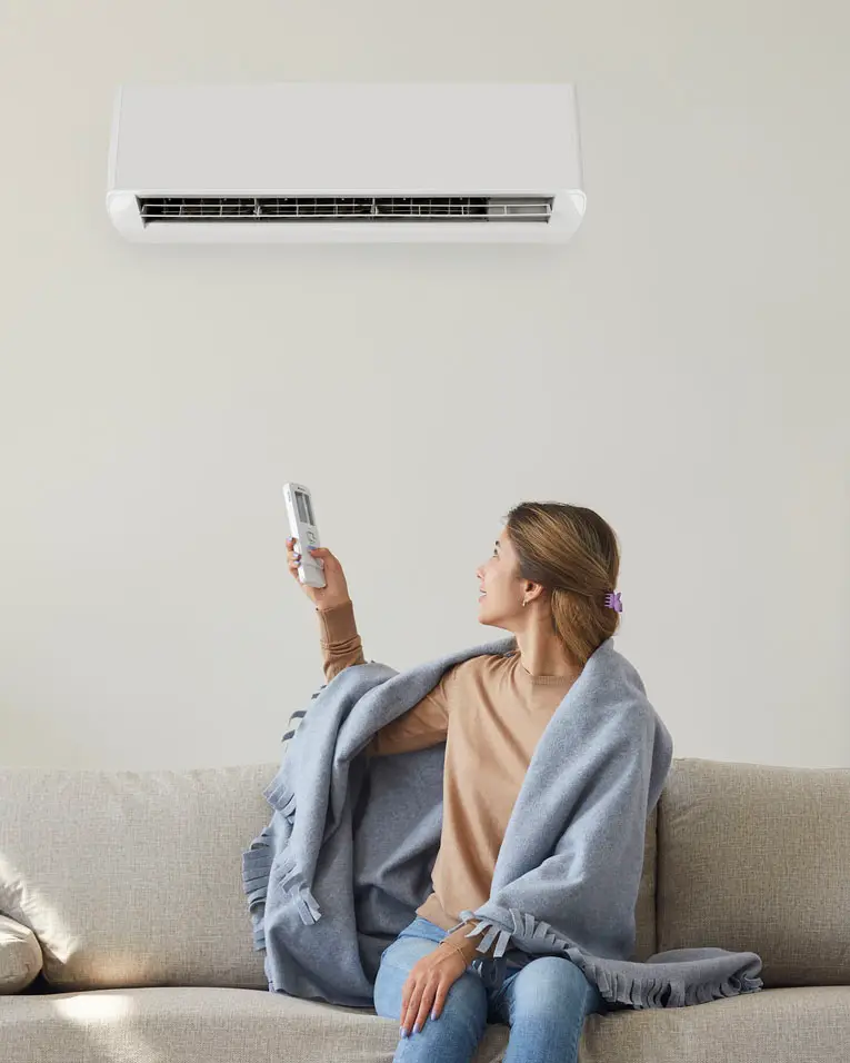 Woman who's sitting on sofa under warm plaid in living room switches off her air conditioner on wall. Young girl adjusting modern AC system, regulating temperature and enjoying cool fresh air at home