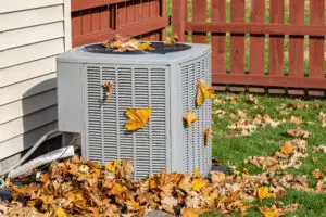 Dirty air conditioning unit covered in leaves during autumn. Home air conditioning, HVAC, repair, service, fall cleaning and maintenance. - in a yard with a fence in the background